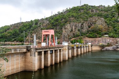 Puerto Pena, Badajoz - İspanya - 10-08-2024: Büyük bir beton baraj, orman kayaları ve dik tepelerle kaplı sakin bir nehri kaplıyor. Barajın birkaç büyük sütunları ve kırmızı bir yapısı var..