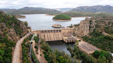 Puerto Pena, Badajoz - İspanya - 10-08-2024: Tepeler ve barajlarla çevrili, yolları, teknik tesisleri ve bir nehri olan büyük bir beton barajın havadan görünüşü.