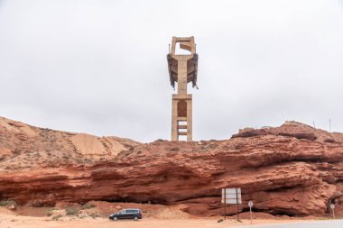 Sidi Ifni, Guelmim-Oued Noun - Fas - 08-08-2024: Bulutlu bir gökyüzüne karşı tek bir beton köprü segmenti yükseliyor, yakınındaki bir araç ve yol işaretleriyle kayalık kırmızı arazide geçiyor.