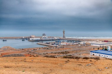 Sidi Ifni, Guelmim-Oued Noun - Fas - 08-08-2024: Bulutlu gökyüzü altındaki deniz kenarı manzarası, binaları, dalgakıranları, havzadaki gemileri ve sanayi yapılarını gösteriyor.