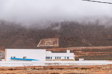 Sidi Ifni, Guelmim-Oued Noun - Fas - 08-08-2024: Beyaz yazıtlı ve Arapça sembollü bir dağ, önplanda, bulutların ve puslu tepelerin altında, modern beyaz çatılı binalar.