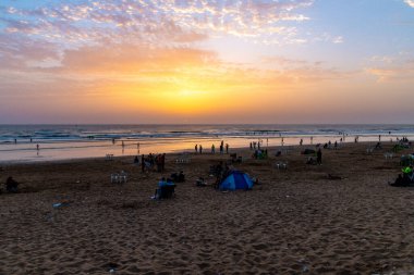 Sidi Ouassay, Chtouka Ait Baha - Fas - 08-09-2024: Gün batımında turuncu ışıkta kumsal, insanlar gruplar halinde, sandalyeler, çadırlar ve sakin dalgalar halinde parçalı bulutlu bir gökyüzü altında.