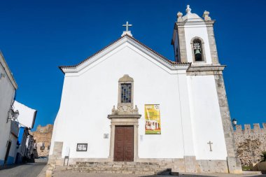 Sines, Alentejo - Portekiz - 09-23-2024: Gable çatı ve çan kulesine sahip beyaz kilise; renkli bir poster ve mavi gökyüzü altında bitişik binalar ile taştan çerçevelenmiş portal.