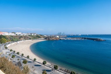 Sines, Alentejo - Portekiz - 09-23-2024: Temiz suyla kıvrımlı kumlu körfez; gezinti güvertesi, beyaz binalar, marina ve turnalar mavi, bulutsuz bir gökyüzü altında görünür.