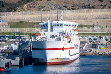 Tanger Med, Fahs-Anjra - Morocco - 08-22-2024: Modern ferry with red and white colors docked in a harbor; surrounded by passenger bridges, loading facilities, and steep hills. clipart