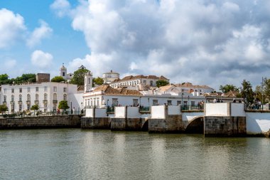 Tavira, Algarve - Portekiz - 09-18-2024: bulutlu bir gökyüzünün altında sakin bir nehre bakan, serin bir şehir manzarasını yansıtan kiremit kaplı binalar..