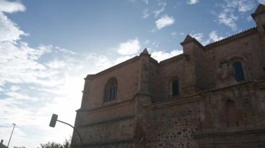 Kilisenin ön cephesi ve çan kulesi, karışık taşlar, kemerli pencereler, dekoratif bir kapı, çanlar, haç ve leylek yuvası..