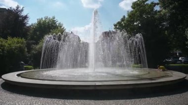 Arona, Piemont - İtalya - 07-04-2024: Kemerli su fıskiyeleri, merkezi sprey, taş kenarı, park ağaçları, mavi gökyüzü ve yakınlarda yürüyen insanlarla çevrili büyük dairesel çeşme.