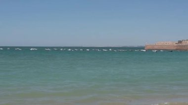 Cadiz, Cadiz - İspanya - 05-07-2024: La Caleta Körfezi 'ne giden Atlantik Okyanusu' nun panoramik manzarası, kıyıya giden küçük tekneler, palmiye ağaçları, beyaz binalar ve bir cankurtaran kulesi.
