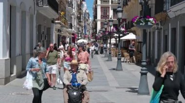 Cadiz, Cadiz - İspanya - 05-07-2024: Cadiz 'deki alışveriş caddesinde kameralara, kafelere ve dükkanlara doğru yürüyen turistler ve sokak lambalarında çiçek saksıları.