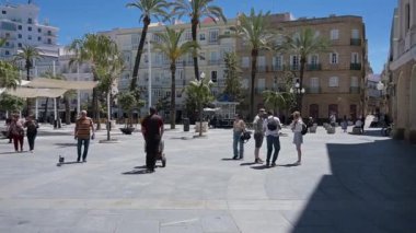 Cadiz, Cadiz - İspanya - 05-07-2024: Restoranlar, palmiye ağaçları, bilet gişeleri, gölgeli yollar ve meydandan geçen bir grup insanın bulunduğu işlek güneşli bir meydanda çevirin.