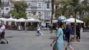 Cadiz, Cadiz - İspanya - 05-07-2024: Güneşli, yoğun bir meydanda insanlarla, restoranlarla, şemsiyelerle, bilet gişeleriyle, gölgeli yollarla, palmiye ağaçlarıyla çevrili binalarla çevrili.