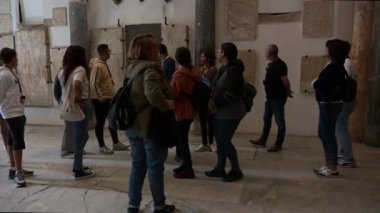 Cordoba, Cordoba - İspanya - 04-08-2024: Kulaklık takmış bir kadın rehber, arkasında taş levhalar olan bir gruba bakıyor, turistler konuşmaya devam ederken turistler arkasını dönüyor.