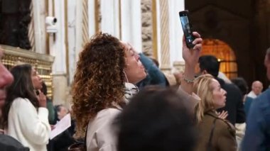 Cordoba, Cordoba - İspanya - 04-08-2024: Mezquitas 'ın içini akıllı bir telefonla kaydeden, kulaklık takan, hareket eden ve izleyen insanlarla çevrili bir kadının yakın çekimi.