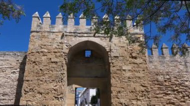 Parlak mavi bir gökyüzünün altında, ön planda ağaç yaprakları ile çerçevelenmiş siperli Cordoba şehir kapısı görüntüsü.