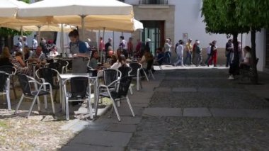 Cordoba, Cordoba - İspanya - 04-09-2024: İnsanlar güneşli, asfalt bir meydanda beyaz şemsiyeler altında yemek yiyor. Turistler arka planda yürürken, bir garson masaları topluyor..