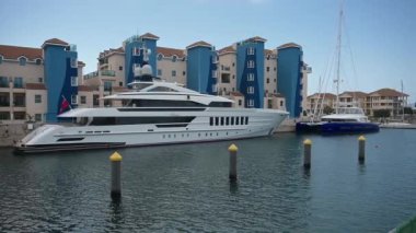 Cebelitars, BOTC - BOTC - 06-03-2024: Cebelitars marinasına yanaşmış lüks yatlar, modern mavi ve bej binalarla çevrili, berrak bir gökyüzünün altında sakin sulardan yansıyan