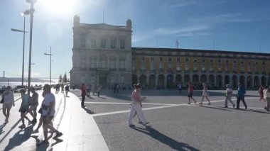 Lissabon, Lisboa - Portekiz - 09-30-2024: Tarihi binalar, yürüyen insanlar ve uzun gölgeler bırakan sokak lambaları olan güneşli bir meydan. Arka planda bir nehir, köprü ve arabalar görünüyor..