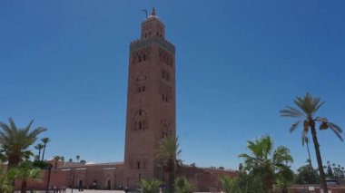 Marrakesch, Marrakesch-Safi - Fas - 08-14-2024: Marakeş 'teki Koutoubia Camii manzarası, yüksek minaresi, etrafındaki palmiye ağaçları ve açık mavi gökyüzü altında komşu bahçeler.