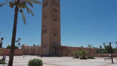Marrakesch, Marrakesch-Safi - Fas - 08-14-2024: Marakeş 'teki Koutoubia Camii manzarası, yüksek minaresi, etrafındaki palmiye ağaçları ve açık mavi gökyüzü altında komşu bahçeler.
