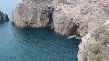 Bir kamera okyanustan kırmızı tepeli beyaz bir deniz fenerine kayalık oluşumlar, yeşillik ve aşağıdaki engin denizi gösterir..