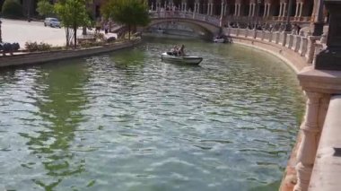Sevilla, Sevilla - İspanya - 07-02-2024: Sevilla 'daki Plaza de Espana' nın sakin kanal suyunda küçük bir tekne dönüyor, arka planda süslü korkuluklar, ağaçlar ve köprü bulunuyor.