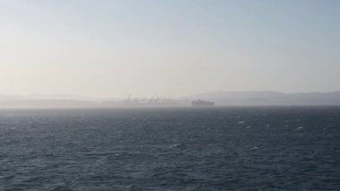 Fas 'a doğru giden bir gemiden bakıldığında endüstriyel vinçler, bir kargo gemisi ve bulutlu, parlak bir gökyüzünün altındaki uzak tepeler görülüyor..