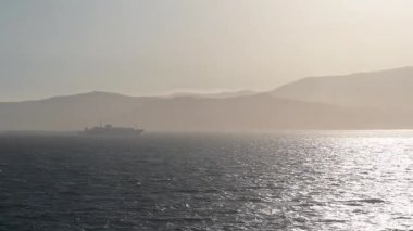 Bir feribot, arka planda Fas kıyı şeridi ile birlikte, altın güneş ışığı altındaki bir gemiden izlenen, ışıldayan su üzerinde yelken açar..