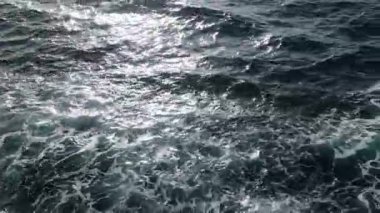 Gemi tarafından üretilen dalgaların yan görüntüsü, parıldayan güneş ışığı karanlık suya yansıyor ve beyaz köpüklü desenler dinamik dalgalanmalar oluşturuyor..