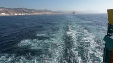 Feribot manzarası, kıyı tepeleri, liman altyapısı ve açık bir gökyüzü ile çevrili beyaz köpüklü suyun okyanusa uzanmasıyla uyanır..