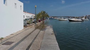 Tavira, Algarve - Portekiz - 09-18-2024: Sel basmış bir nehir bir şehir içinden akar; su gezinti alanının üzerinden yola taşar; park edilmiş arabalar, palmiye ağaçları, demirlemiş botlar ve yoğun bir köprü.