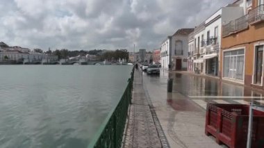 Tavira, Algarve - Portekiz - 09-18-2024: Bir nehir kıyılarını yaya geçidine taşır ve bitişik yolu sular altında bırakır; yayalar suda yürür; park edilmiş arabalar; taş köprü görünür.