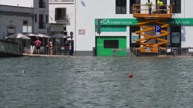 Tavira, Algarve - Portekiz - 09-18-2024: Sonsuzluk havuzunu andıran sel basmış bir nehrin karşısına bakın; su karşı sokağı sular altında bırakıyor; bisikletçi ve işçiler beyaz bir binanın yanındaki asansörde duruyorlar.