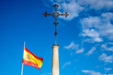 Üzerinde kavisli desenler olan süslü bir metal haç, üzerinde açık mavi bir gökyüzünün altında dalgalanan bir İspanyol bayrağı bulunan uzun bir taş sütunun üzerinde duruyor..