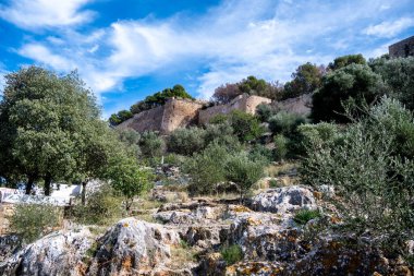 Ağaçlar ve çalılarla çevrili eski bir taş kale duvarı kayalık arazinin üzerinde yükseliyor. Ön planda düz olmayan kayalar, çimenler, yosunlar ve bulutlu mavi bir gökyüzü var..