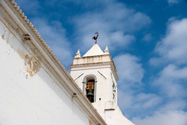 Üzerinde hava durumu horozu olan beyaz bir kilisenin çan kulesine yakın çekim, açık mavi gökyüzüne karşı kurulmuş..