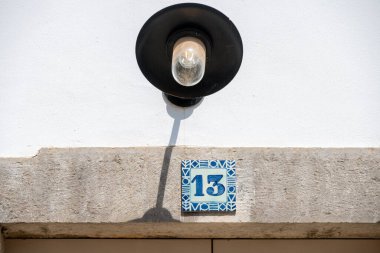 Close-up of a house number plate with the number 13 in blue ceramic pattern, above it a black outdoor lamp on a white wall. clipart