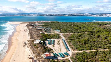 Geniş bir kumsal, yeşil kum tepeleri ve mavi gökyüzünün altındaki Lissabon ile geniş bir kıyı şeridinin panoramik görüntüsü.