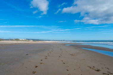Ayak izleri ve su birikintileriyle geniş kumlu bir sahil. Arka planda dağınık bulutlar, binalar ve aylaklar olan mavi gökyüzü..