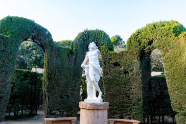 Bir kadın figürü klasik mermer heykel yuvarlak bir kaide üzerinde durur, uzun kesilmiş çitlerle çevrili yapılandırılmış bir bahçede kemerli açıklıklar ile.