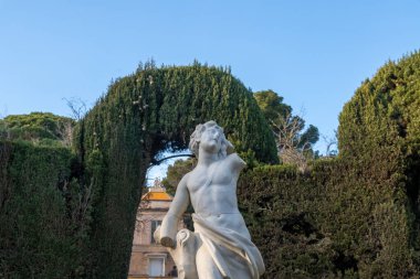 Yıpranmış mermer bir kadın heykeli, kolları olmayan bir kadın heykeli, ağaçlarla kaplı, düzgün kesilmiş bir çit kemerinin önünde duruyor..