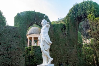 Bir kaidedeki klasik mermer heykel, arka planda kubbeli bir pavyon ve yapılandırılmış bir bahçedeki budanmış çit kemerleri arasında duruyor..