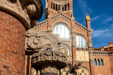 Ön planda oyma detaylar olan dekoratif taş bir sütun. Arkasında da Sant Pau hastanesinin süslü kulesi var..