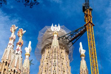 Close-up of Sagrada Familia tower under construction, wrapped in scaffolding, with intricate details and a crane extending against a clear blue sky. clipart