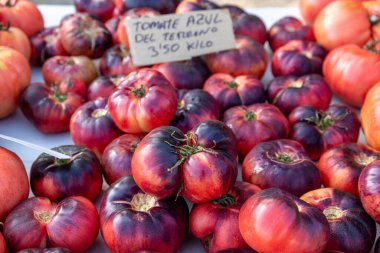 Denia, Alicante - Spain - 12-01-2024: A pile of heirloom tomatoes with a unique dark red and purple hue on a market table, with a handwritten price sign in the background. clipart