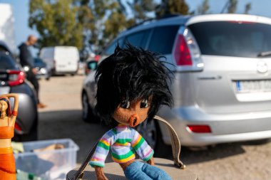 Denia, Alicante - Spain - 12-01-2024: A handmade fabric doll with black shaggy hair, a colorful striped sweater, and denim pants, displayed on a market table with cars in the background. clipart