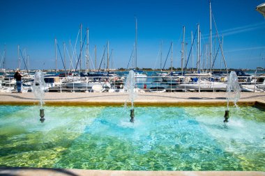 A fountain with four jets of clear water in front of a sailboat harbor, surrounded by pathways and a bright blue sky. clipart