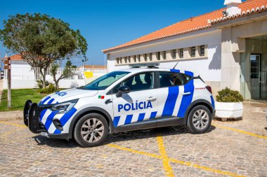 A white-blue Portuguese police car parked on a cobblestone lot, surrounded by modern buildings and trees in the background. clipart