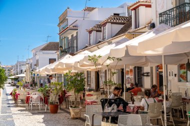 A street cafe with white tables and umbrellas, with guests enjoying food and drinks, and white buildings with red roofs in the background. clipart