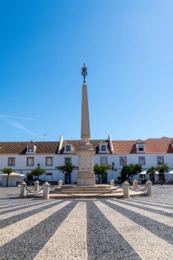 Asfaltlı bir meydanda duran büyük bir dikilitaş, kırmızı çatıları ve üzerinde açık mavi bir gökyüzü olan beyaz binalarla çevrili..